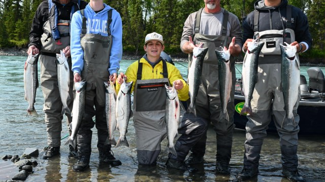 Five people in waders holding fish by a river with trees in the background.