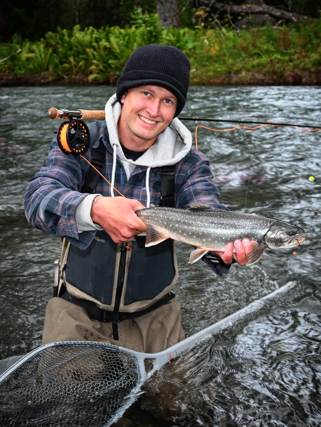 Person in waders holding a fish and smiling in a river with a fly fishing rod and net.