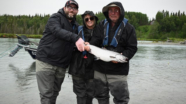 Three people wearing fishing gear holding a fish by a river with trees in the background.